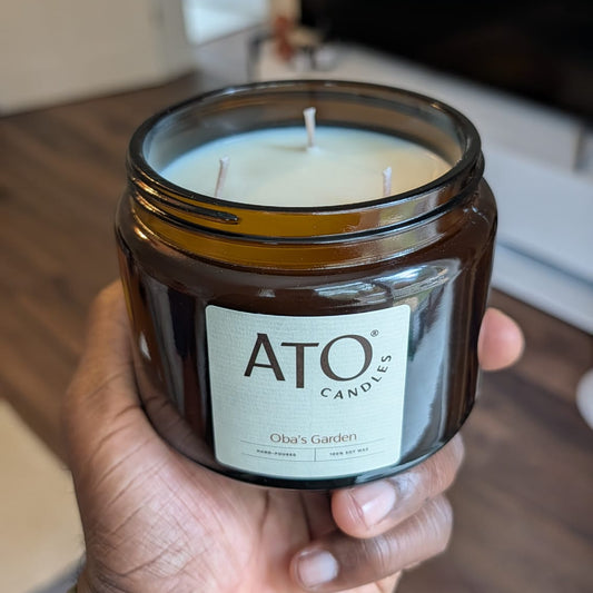 A hand holding a brown glass jar candle with a white label that reads 'ATO CANDLES' and 'Olor de Garden' in a living room setting.